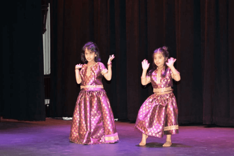 Two young girls dressed in traditional Indian dance costumes perform on stage, their expressions and hand gestures showing concentration and joy during a kids dance performance.