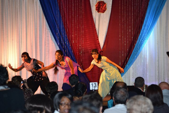 Three dancers performing on a stage decorated with red, white, and blue drapes, in front of an audience. The dancers are captured mid-movement, showcasing their expressive dance styles at what appears to be a community event or celebration.
