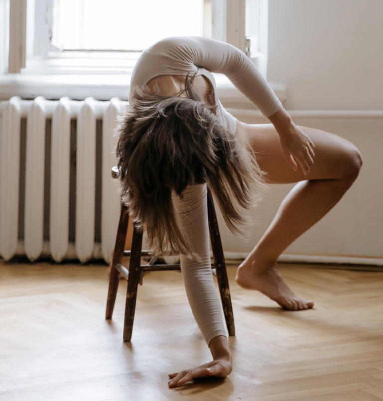 A dancer in a neutral-toned bodysuit performs an intricate pose on a wooden chair, demonstrating flexibility and strength in a bright room with a vintage radiator and window providing natural light. Her movement and form are the focus of this artistic dance practice setting.
