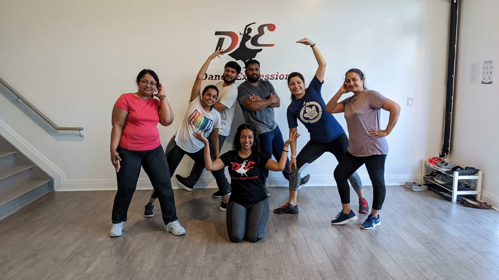 A group of adults posing with playful expressions and dance-inspired gestures in Dance Expression Studio, wearing casual workout clothes and sneakers. Behind them, the wall features the Dance Expression logo, indicating the lively and inclusive atmosphere of the dance class they are attending.