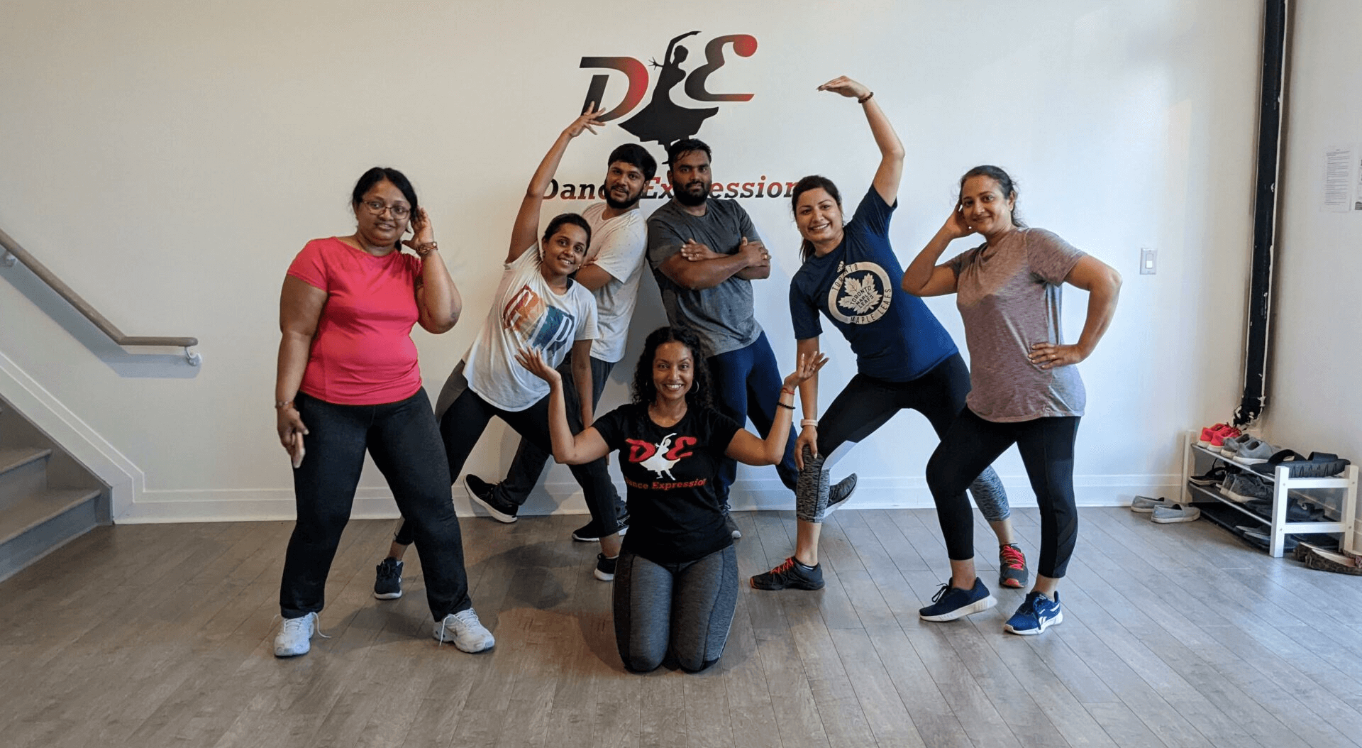 A group of adults posing with playful expressions and dance-inspired gestures in Dance Expression Studio, wearing casual workout clothes and sneakers. Behind them, the wall features the Dance Expression logo, indicating the lively and inclusive atmosphere of the dance class they are attending.