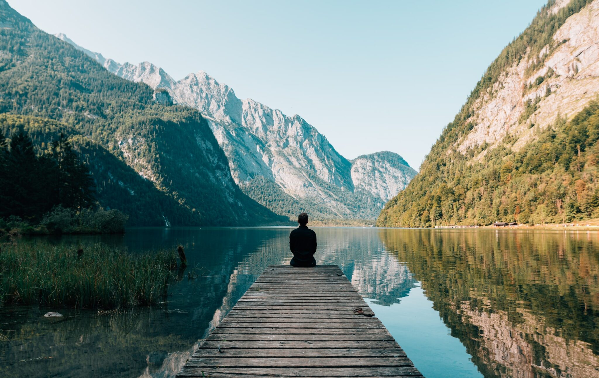 A person sitting peacefully at the end of a wooden dock, overlooking a serene mountain lake with reflections of the surrounding forested hills on the water's surface, under a clear sky, creating a tranquil and picturesque natural setting.