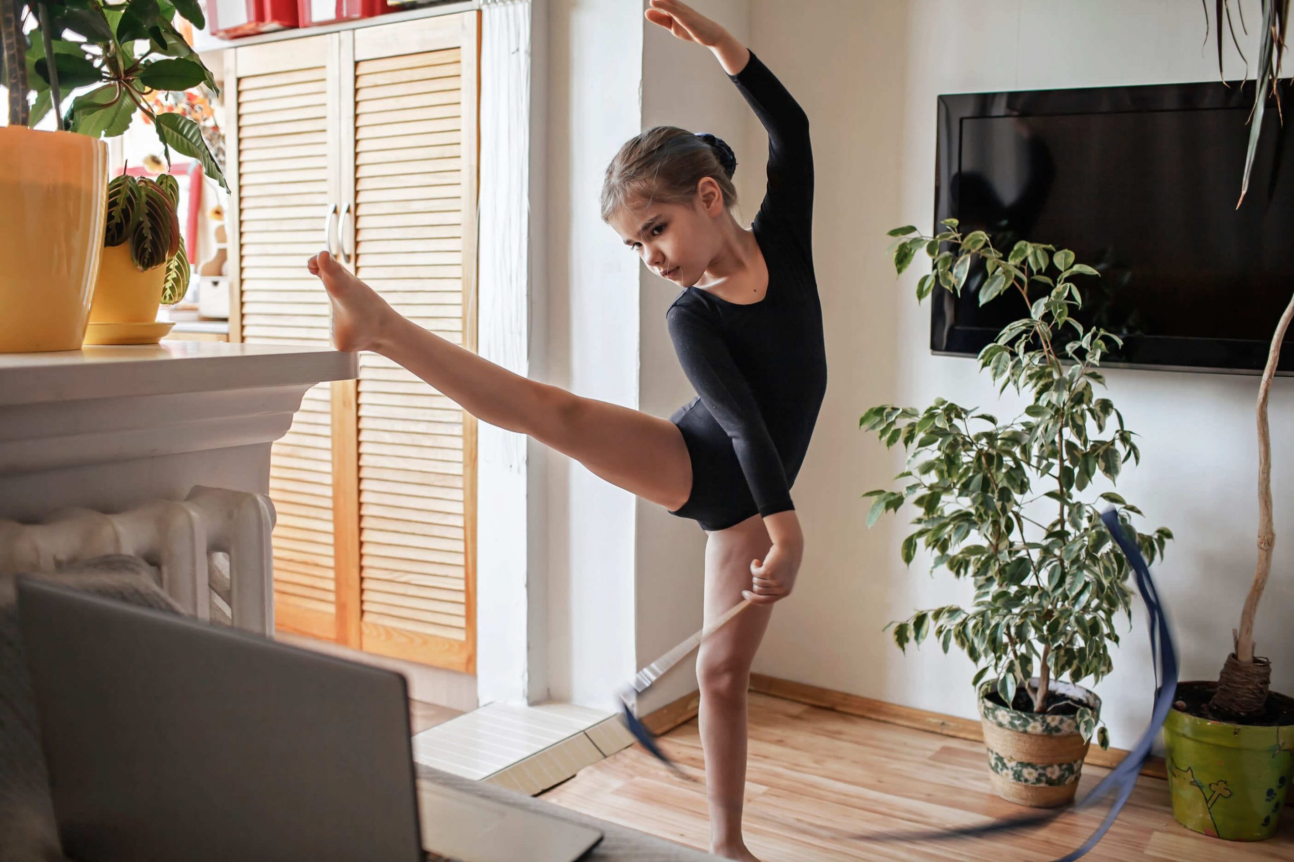 A young ballet dancer practicing at home, exhibiting poise and flexibility with a high leg extension. The room is bright and cozy, with houseplants and a TV in the background, indicative of a comfortable, personal space adapted for dance practice.