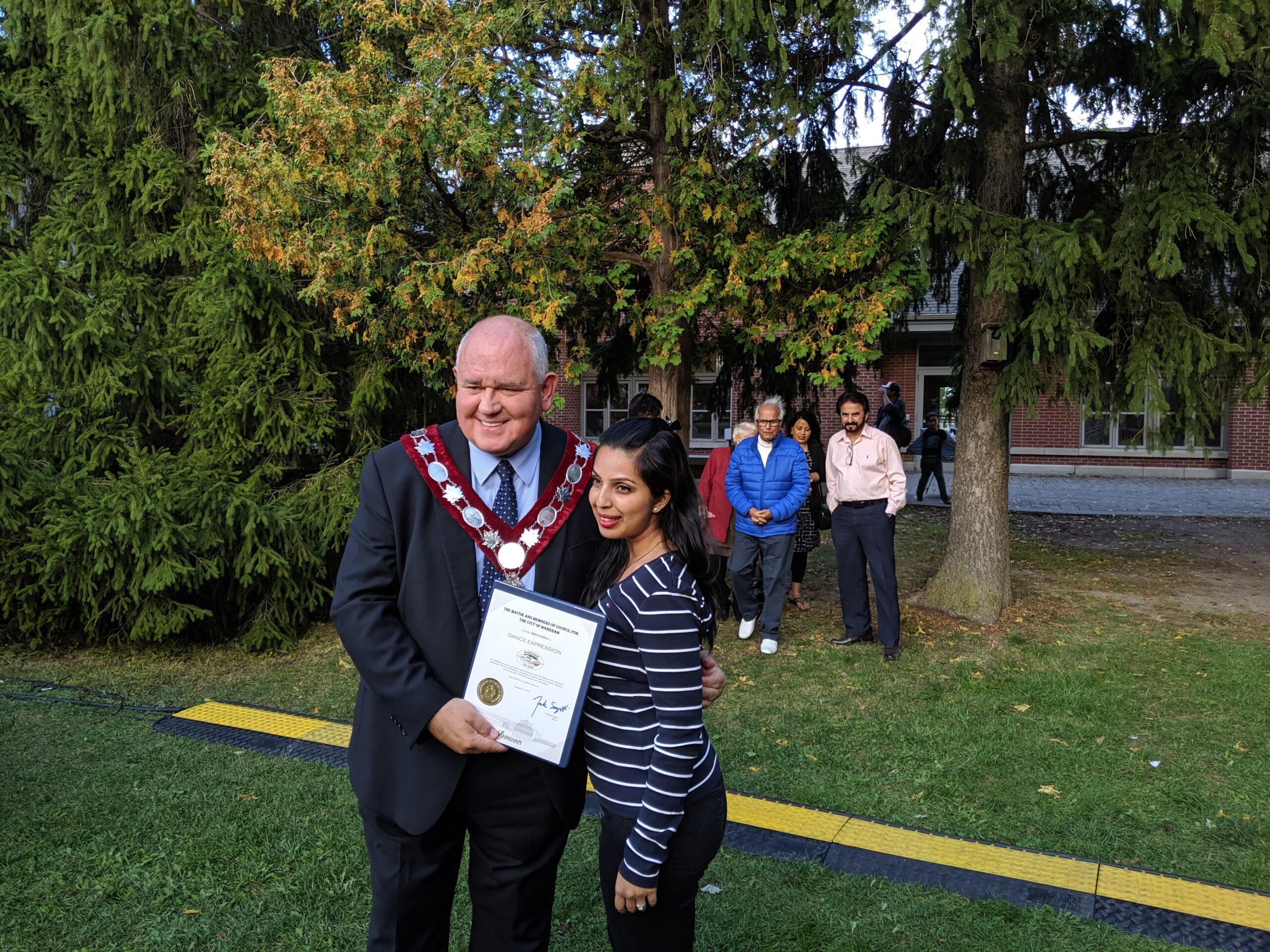 Mayor of Markham, Frank Scarpitti, in ceremonial chain and Creative director and founder of Dance Expression Studio, holding a certificate posing for a photograph outdoors, with observers in the background, at the Markham 225th anniversary.