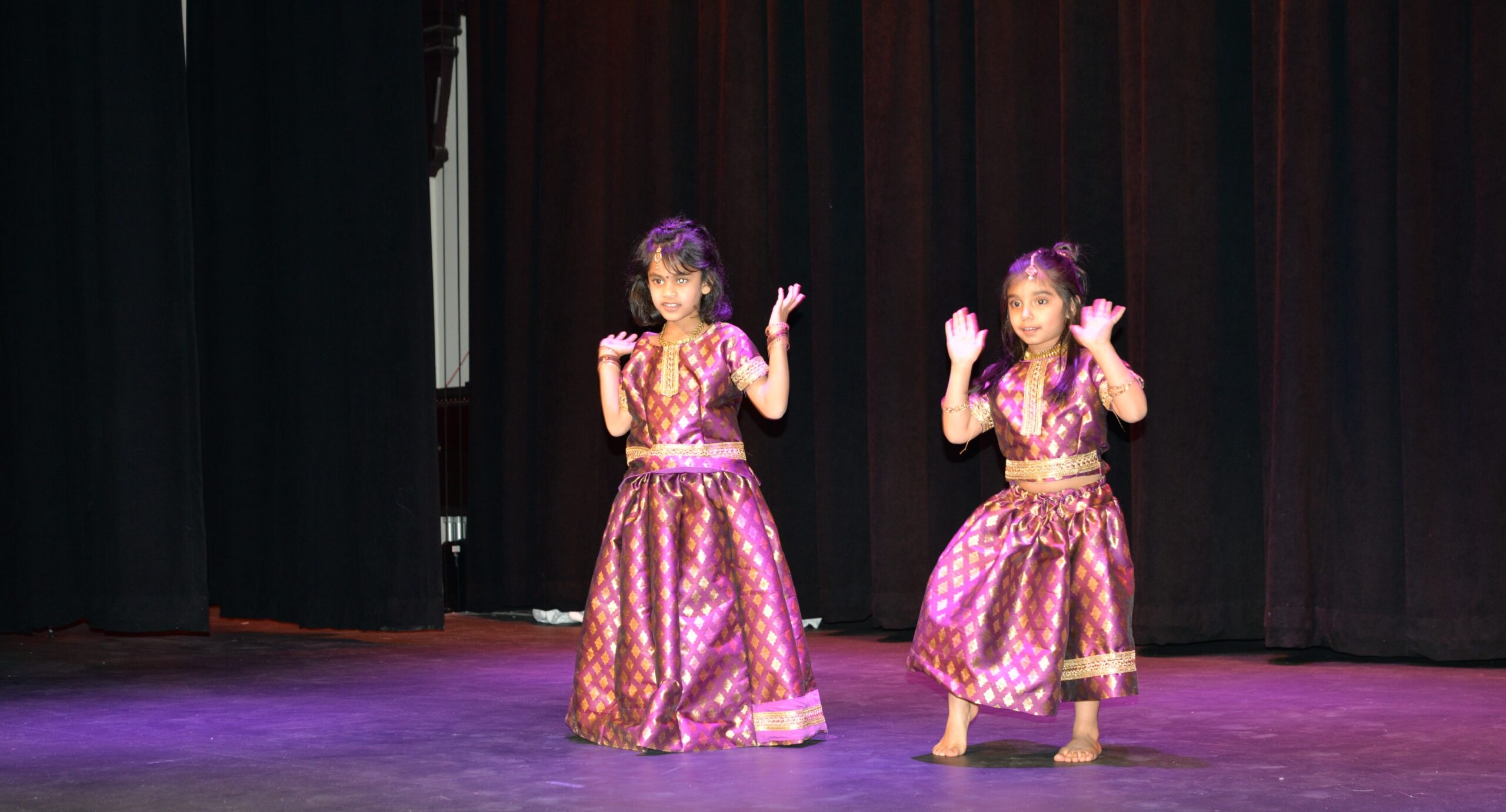 Two young girls dressed in traditional Indian dance costumes perform on stage, their expressions and hand gestures showing concentration and joy during a kids dance performance.