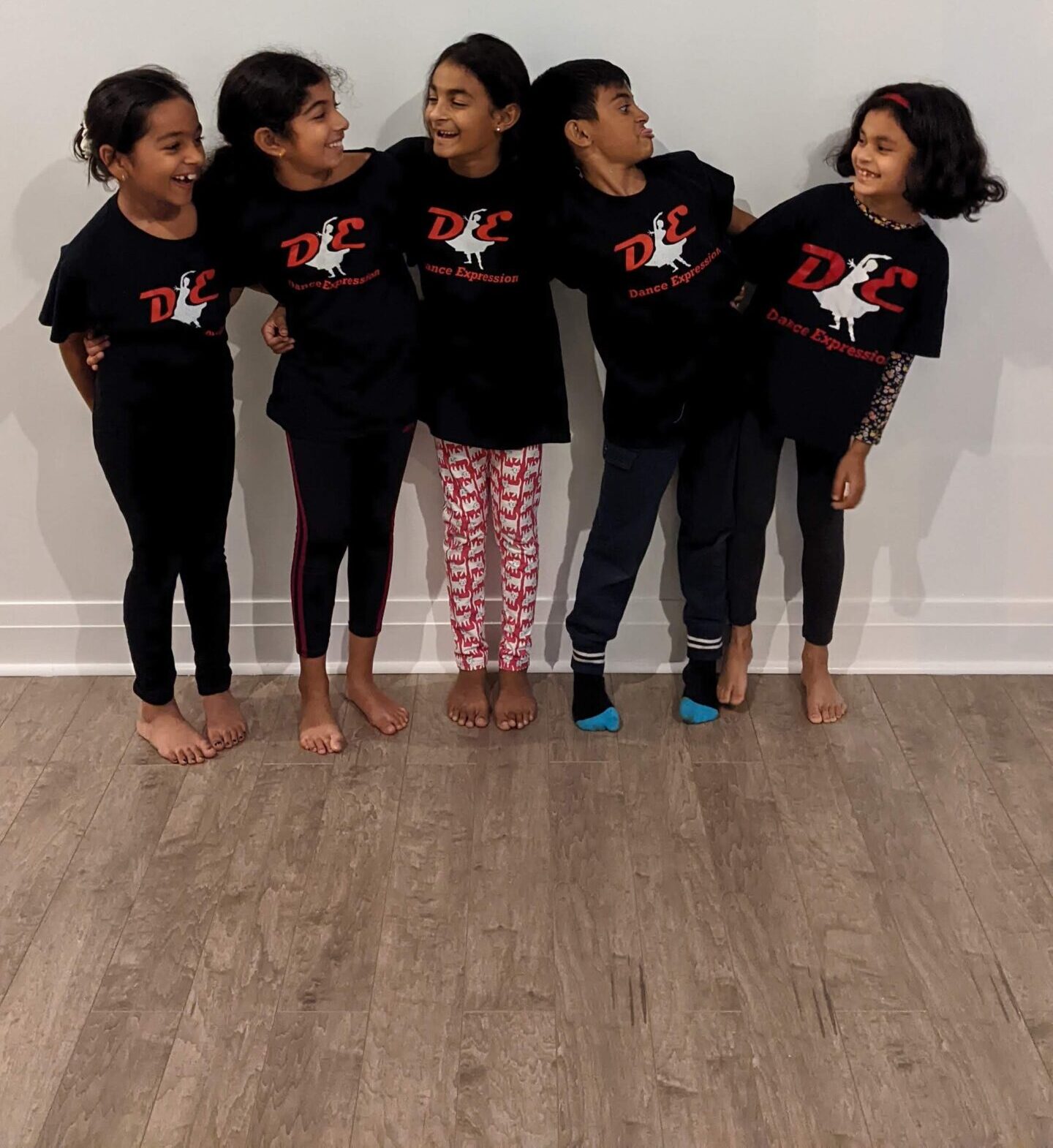 Five smiling children wearing black Dance Expression t-shirts standing in a row, looking at each other and sharing a joyful moment together, possibly after a dance class at the Dance Expression studio. They are barefoot, indicating a casual and comfortable environment conducive to dance practice.