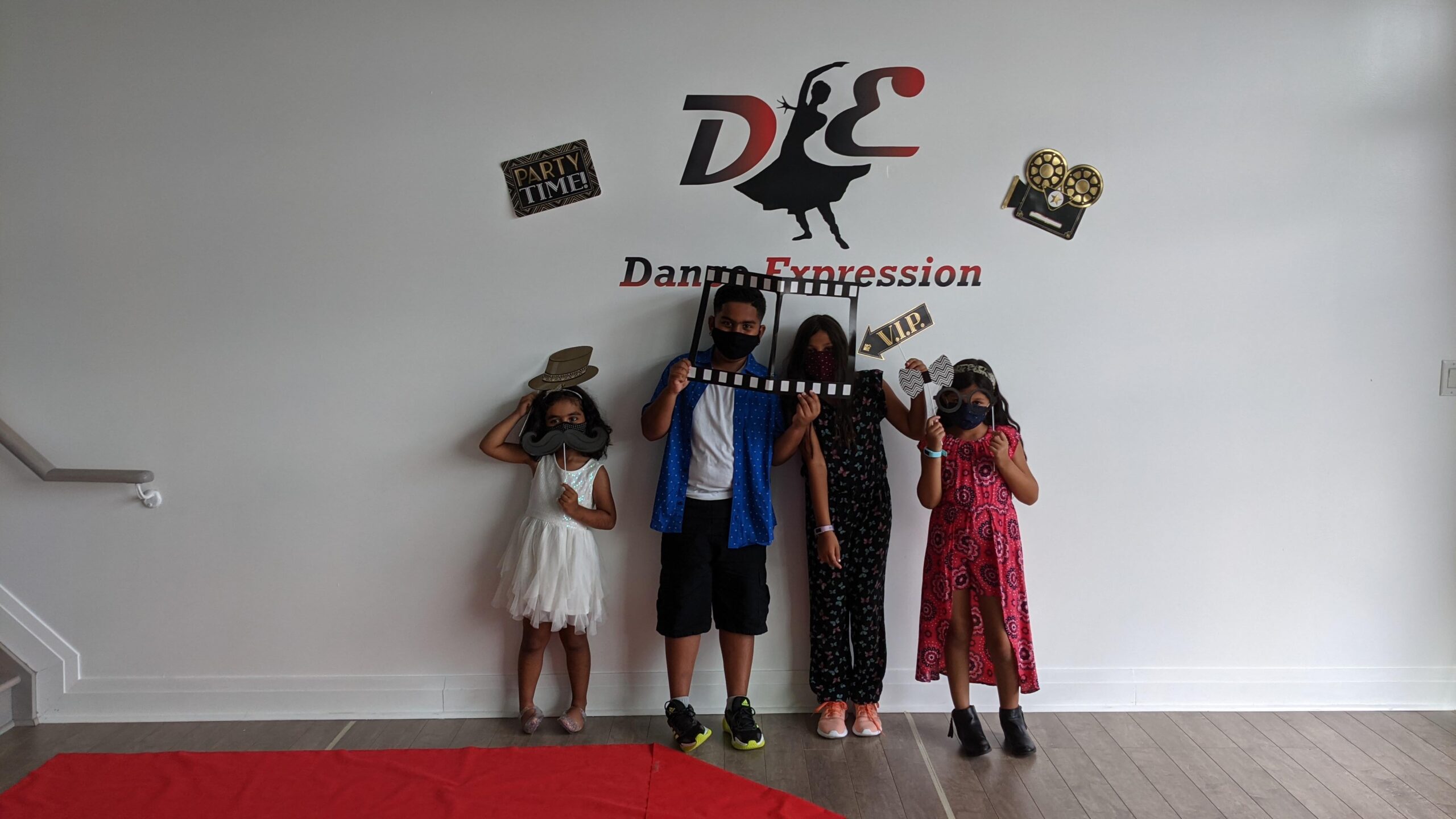 Four individuals are playfully posing with photo props in front of a Dance Expression studio wall, decked out in festive decorations, showcasing the enjoyable times they experienced at the Dance Expression Summer Camp program.