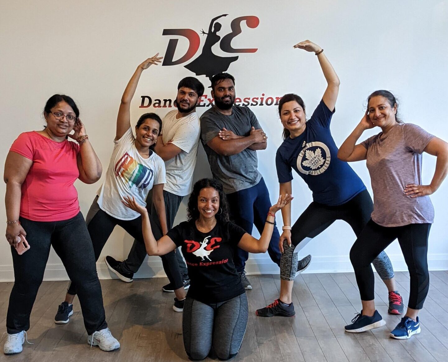 A group of adults posing with playful expressions and dance-inspired gestures in Dance Expression Studio, wearing casual workout clothes and sneakers. Behind them, the wall features the Dance Expression logo, indicating the lively and inclusive atmosphere of the dance class they are attending.